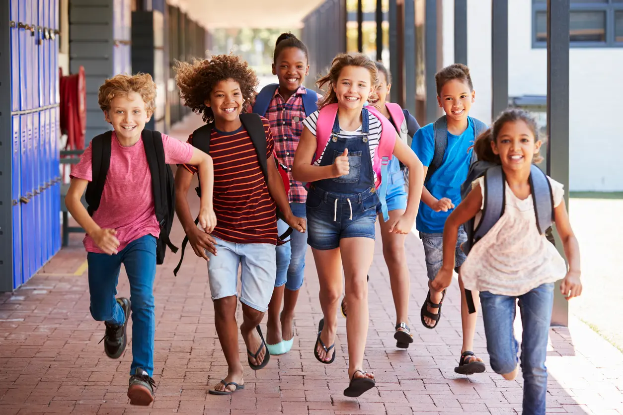 A group of six joyful children with backpacks runs down a school corridor, smiling and laughing. Their energetic movement conveys excitement and happiness.
